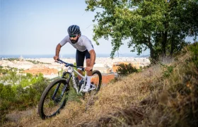 Una persona que lleva casco y gafas de sol pedalea cuesta arriba en una bicicleta de montaña equipada con ruedas FSA por un sendero cubierto de hierba, disfrutando de un control total mientras la ciudad y el mar se extienden al fondo.