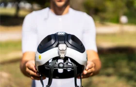 Una persona con camisa blanca sostiene un casco de bicicleta polivalente POC Amidal en blanco y negro, con una luz trasera incorporada, al aire libre en un día soleado: ideal tanto para aventuras por carretera como por grava.