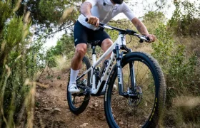 Una persona que lleva casco, gafas de sol y equipación de ciclismo circula en una bicicleta de montaña con ruedas FSA KFX i30 por un sendero de tierra rodeado de hierba y árboles, disfrutando de un control total sobre el terreno accidentado.