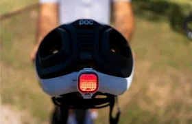 Primer plano de un casco de bicicleta polivalente con una luz trasera de seguridad roja acoplada, sostenido por una persona que se encuentra al aire libre en un día soleado; ideal tanto para aventuras en carretera como en grava.