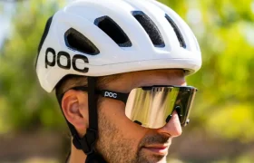Una persona que lleva un casco de ciclismo polivalente blanco de la marca POC, gafas de sol y un maillot de ciclismo de color claro está de pie al aire libre, con árboles al fondo, lista para una aventura por carretera o por rutas de grava.