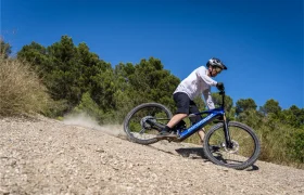 Una persona con casco baja en bicicleta de montaña azul por un sendero rocoso, rodeada de árboles y bajo un cielo azul y despejado. Gracias al Bosch Performance Upgrade, disfruta de una mayor potencia y control mientras el polvo se levanta tras la rueda trasera.