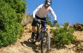 Una persona que lleva casco y gafas de sol recorre en bicicleta de montaña, equipada con un sistema Bosch Performance Upgrade, un sendero de tierra rocoso rodeado de matorrales verdes bajo un cielo azul y despejado, disfrutando de más potencia y más control en cada curva.