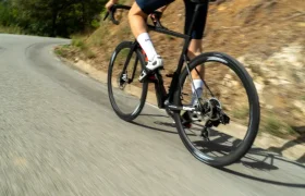 Un ciclista, con casco blanco y ropa azul, pedalea cuesta arriba en una bicicleta de carretera equipada con neumáticos Schwalbe Pro One en un día soleado, con árboles y un camino en pendiente al fondo.