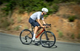 Un ciclista que lleva un casco blanco y un maillot gris pedalea en una bicicleta de carretera negra y roja equipada con neumáticos Schwalbe Pro One Aero tanto en la rueda delantera como en la trasera, deslizándose por un camino asfaltado con vegetación al fondo.