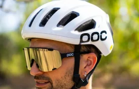 Una persona que lleva un casco de bicicleta blanco «casco polivalente» y unas grandes gafas de sol de espejo está de pie al aire libre, mirando hacia un lado, lista para una aventura por carretera o por rutas de grava.
