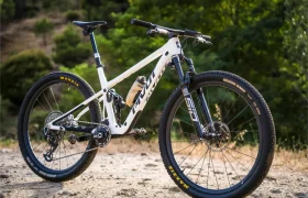 Una bicicleta de montaña blanca con suspensión total, equipada con neumáticos Maxxis y ruedas FSA KFX i30, está aparcada en un camino de tierra rodeado de árboles frondosos, lo que ofrece a los ciclistas un control total incluso en los terrenos más exigentes.