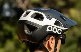 Se ve desde atrás a una persona que lleva un casco de bicicleta POC Cularis Pure, en blanco y negro, con viseras para protegerse del sol, de pie al aire libre con árboles al fondo.