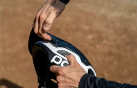 Una persona se ajusta la visera de un casco de ciclista POC Cularis Pure, en blanco y negro, al aire libre, sujetando con las manos los puntos de fijación de la visera bajo el cálido sol.