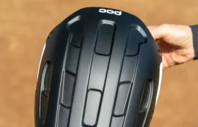 Una mano sostiene un casco de ciclismo POC Cularis Pure negro con acabado mate y ranuras de ventilación verticales, diseñado para adaptarse al clima, al aire libre en un camino de tierra.