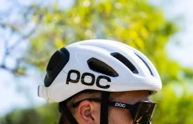 Persona que lleva un casco de ciclismo polivalente blanco de la marca POC y gafas de sol negras al aire libre, con follaje verde al fondo.