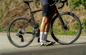 Un ciclista vestido de negro está de pie junto a una bicicleta de carretera negra equipada con neumáticos Schwalbe Pro One en una carretera asfaltada, con un pie en el suelo y el otro en un pedal.
