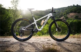 Una bicicleta de montaña blanca con suspensión total, equipada con ruedas FSA KFX i30 y neumáticos negros, está aparcada en un camino de tierra, ofreciendo un control total mientras descansa entre los árboles y las pintorescas colinas que se divisan al fondo.