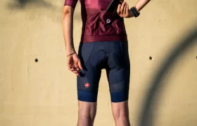 Un ciclista con maillot granate, pantalones cortos tipo culotte A/C azul marino, calcetines blancos y casco está de pie frente a una pared, guardándose el móvil en el bolsillo trasero, listo para rendir al máximo gracias al conjunto Castelli Climber’s A/C.