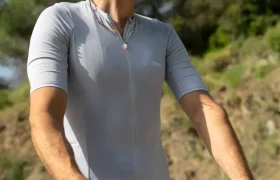 Una persona que lleva casco, gafas de sol y un maillot de ciclismo POC Cadence de color gris claro va en bicicleta al aire libre en un día soleado, con árboles al fondo, disfrutando de la comodidad para pedalear que le proporciona su culotte Cadence Cargo.