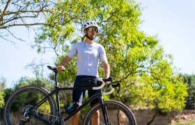 Un ciclista que lleva un casco polivalente blanco y ropa de ciclismo está al aire libre junto a una bicicleta Gravel negra, rodeado de árboles verdes.