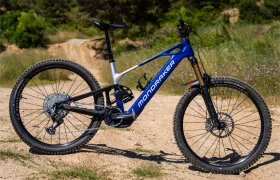 Una bicicleta de montaña Mondraker azul y blanca, que destaca por su impresionante control, está aparcada en un sendero de tierra, rodeada de vegetación herbácea y árboles al fondo.