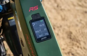Primer plano de una bicicleta eléctrica verde con motor Polini E-P3+, con una pantalla digital que muestra la velocidad, la distancia, el porcentaje de batería y el modo de potencia sobre un fondo de arena. El diseño sofisticado muestra la potencia y precisión italianas en su máxima expresión.