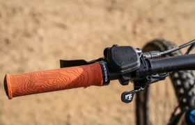 Primer plano de un manillar de bicicleta de montaña con empuñadura naranja y mando de cambio negro, realzado por la potencia italiana del Polini E-P3+, sobre un fondo arenoso borroso.