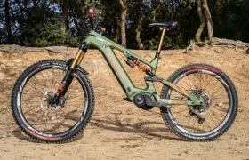 Una bicicleta de montaña eléctrica ecológica con suspensión total, equipada con el potente motor Polini E-P3+ y frenos de disco, está estacionada en un camino de tierra, rodeada de árboles.