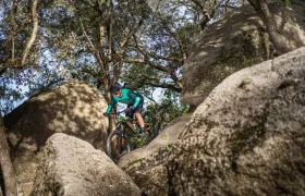 Un ciclista, haciendo gala de la potencia italiana del Polini E-P3+, avanza hábilmente entre grandes rocas en una zona boscosa, vestido con una chaqueta verde y un casco.