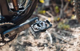 Primer plano de un pedal de bicicleta de montaña, en el que se ven los pedales Shimano DEORE XT SPD Trail acoplados al brazo de la biela, con parte de la cadena y la suspensión trasera visibles en un entorno al aire libre.