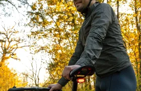 Un ciclista con casco, gafas de sol y ropa oscura con el CLIP SIGMA NYTE colocado se sitúa junto a una bicicleta en un camino forestal bordeado de follaje otoñal.