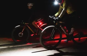 Dos ciclistas circulan de noche por una carretera oscura, iluminados por luces traseras rojas y un faro brillante, evocando el espíritu de aventura de States of Elevation o los legendarios viajes nocturnos de Kilian Jornet.