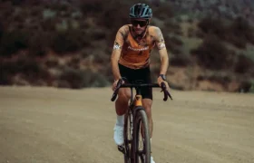 Un ciclista con casco y gafas de sol conduce una bicicleta de grava por un camino de tierra, canalizando el espíritu de States of Elevation - Kilian Jornet, con colinas onduladas y un cielo nublado de fondo.