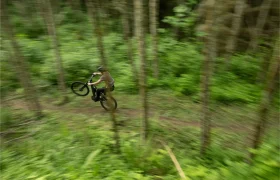Una persona monta en una bicicleta Santa Cruz Vala AL, realizando un caballito en un sendero de tierra a través de una zona verde y boscosa.