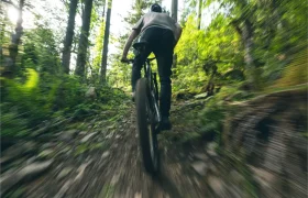 Una persona conduce una bicicleta de montaña Santa Cruz Vala AL por un sendero pedregoso de tierra a través de un bosque denso y verde. La imagen está difuminada para transmitir velocidad y destaca el ligero cuadro de aluminio de la bicicleta.