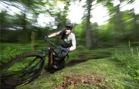 Una persona con casco monta en una bicicleta de montaña Santa Cruz Vala AL, su ligero cuadro de aluminio atraviesa rápidamente un bosque con árboles verdes difuminados al fondo, sugiriendo movimiento.
