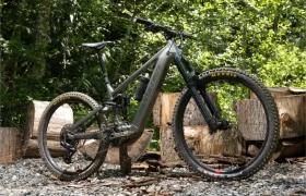 Una bicicleta de montaña Santa Cruz Vala AL con suspensión total, neumáticos anchos y cuadro de aluminio está aparcada sobre grava delante de grandes troncos cortados, rodeada de verde follaje.