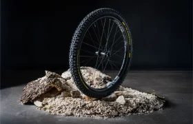 Una rueda de bicicleta de montaña Pirelli Scorpion se yace en posición vertical sobre un montón de rocas, grava y corteza, con un fondo oscuro, lo que pone de manifiesto la gran resistencia de los neumáticos de MTB.