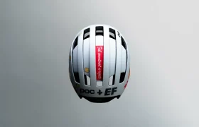 Casco de bicicleta blanco visto desde arriba, con rejillas de ventilación negras, varios logotipos y una etiqueta roja con texto en el centro.