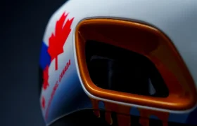 Primer plano de un casco de carreras blanco con una rejilla de ventilación naranja, una hoja de arce roja y la inscripción «CANADA». También se aprecian detalles en azul y rojo en el casco.
