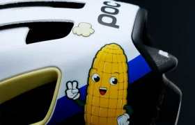 Un casco de bicicleta blanco y negro muestra una mazorca de maíz de dibujos animados con brazos, piernas y una cara sonriente, haciendo el signo de la paz, junto con pequeños dibujos de palomitas.