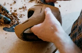 Una persona tallaba los detalles de una máscara de madera con una herramienta manual; sobre la superficie de trabajo había virutas de madera, herramientas de tallado y un casco integral POC.