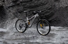 Una bicicleta de montaña Avinox de suspensión total, con cuadro gris y detalles amarillos, está aparcada sobre suelo rocoso húmedo frente a una oscura pared de roca.