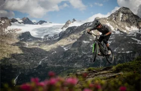Una persona monta en bicicleta de montaña, equipada con neumáticos Pirelli Scorpion MTB, y se lanza desde un saliente rocoso, desafiando la gravedad con montañas nevadas y un glaciar al fondo, y flores silvestres de color rosa en primer plano.