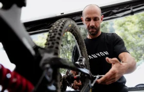 Un hombre que lleva una camiseta negra de Canyon repara una bicicleta de montaña, prestando especial atención a la cadena y los cambios. Equipada con unos robustos neumáticos Pirelli Scorpion para MTB, ideales para senderos de gravedad, su bicicleta destaca sobre el fondo de los árboles y parte de una copa.
