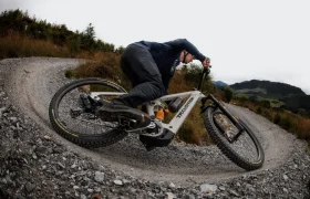 Una persona recorre una curva de grava en un sendero de tierra con una bicicleta de montaña Transition equipada con neumáticos Pirelli Scorpion MTB, rodeada de hierba y colinas lejanas bajo un cielo nublado.