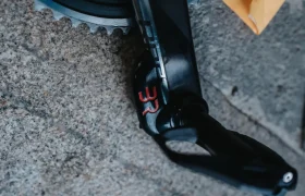 Primer plano de un brazo de biela de bicicleta negro con marcaje rojo "B2" y un potenciómetro Body Rocket integrado, unido a un engranaje, sobre un fondo de piedra texturizada.
