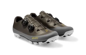 Un par de zapatillas de ciclismo marrones y grises con cierres de dial negros, suelas blancas y el logotipo «QUOC» en el lateral.