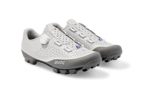 Un par de zapatillas de ciclismo blancas y grises con tacos negros, parte superior perforada y sistema de cierre con rueda giratoria.