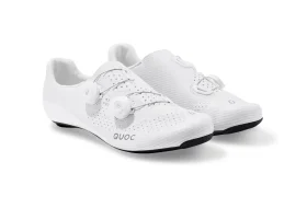 Un par de zapatillas de ciclismo blancas con suela negra, con la parte superior perforada y sistema de cierre con rueda giratoria, colocadas una al lado de la otra sobre un fondo blanco.