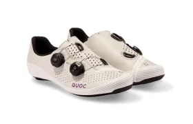 Un par de zapatillas de ciclismo de color beige con cierres negros, parte superior perforada y el nombre de la marca «QUOC» impreso en morado en el lateral.