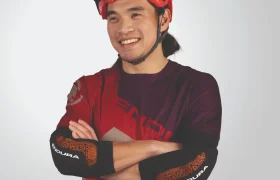 Una persona que lleva un casco rojo y una camiseta de ciclismo Endura, con los brazos cruzados y sonriendo sobre un fondo liso.