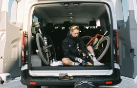 Un ciclista se sienta en la parte trasera de una furgoneta abierta con dos bicicletas en su interior, calzándose las zapatillas de ciclismo. Una bolsa de lona negra descansa en el suelo, como parte de su organizado sistema de viaje.