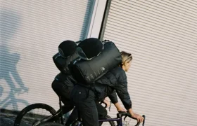 Dos personas con mochilas ASSOS pasan en bicicleta por delante de la puerta cerrada de un garaje industrial en un día soleado, mostrando su sistema de viaje ciclistas.